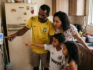 Foto de família brasileira apontando para o medidor de tomada conectado a uma geladeira antiga, com expressão de concentração enquanto leem o display do aparelho